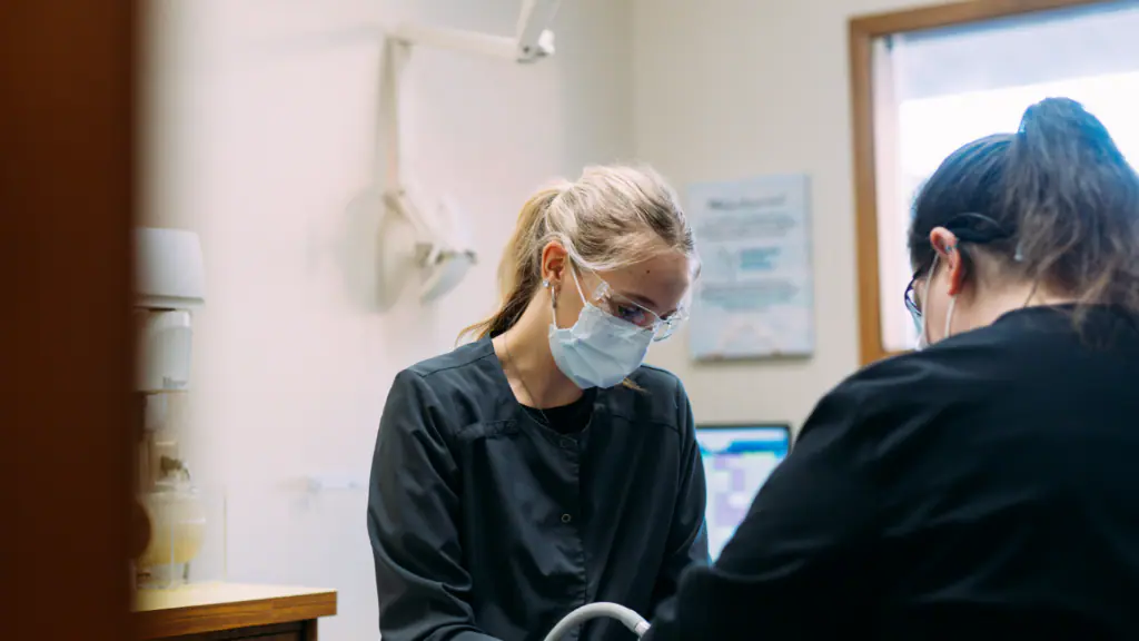 Two healthcare workers in scrubs and masks perform a procedure in a clinic, with equipment visible in the background.