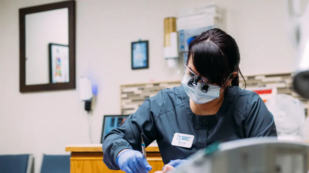 Dental professional in scrubs and mask performing a procedure in a clinical setting wearing magnifying loupes and gloves.