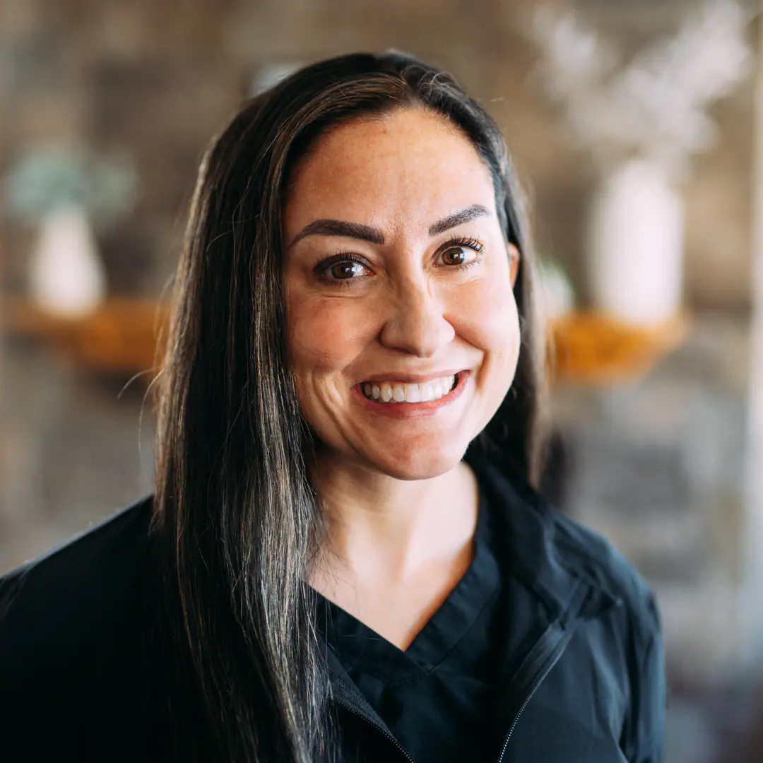 Smiling woman with long dark hair and brown eyes, wearing a black jacket indoors, facing the camera.