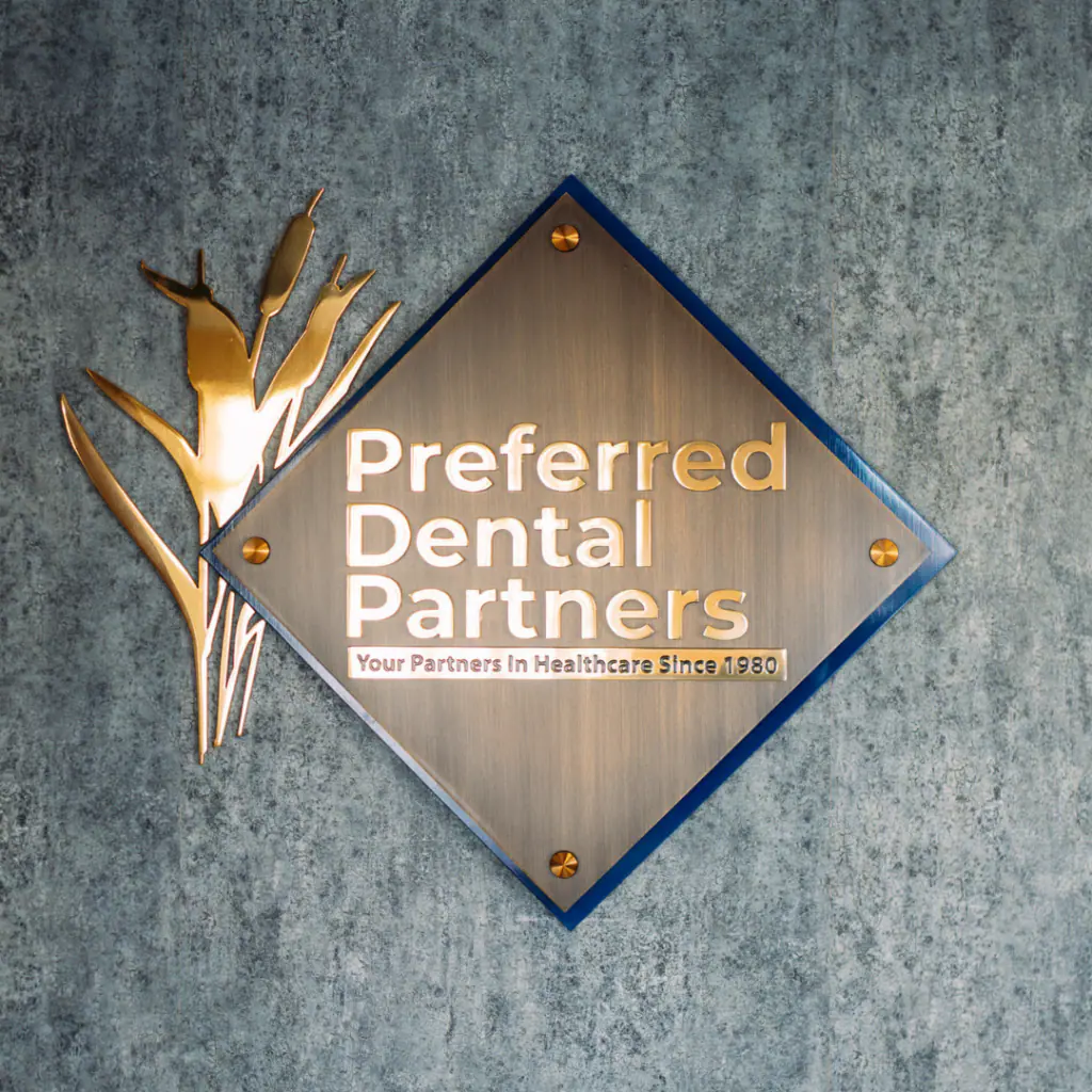 Diamond-shaped metal sign reading "Preferred Dental Partners" with a gold decorative plant on the left, mounted on a textured blue-green wall, including the tagline "Your Partners In Healthcare Since 1980".
