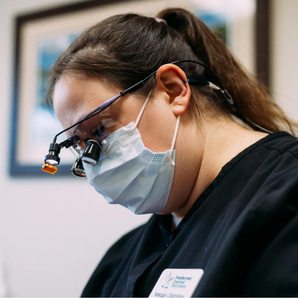 Healthcare worker wearing a protective mask and head-mounted magnifying loupes, focused on a task in a clinical setting.