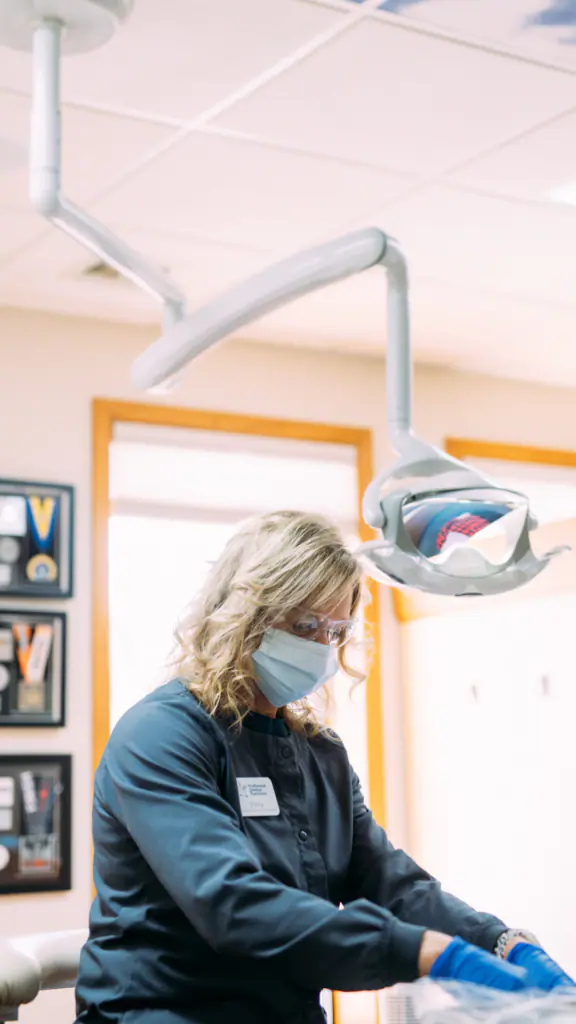 Dental professional in scrubs and mask performing a procedure under an overhead dental light.