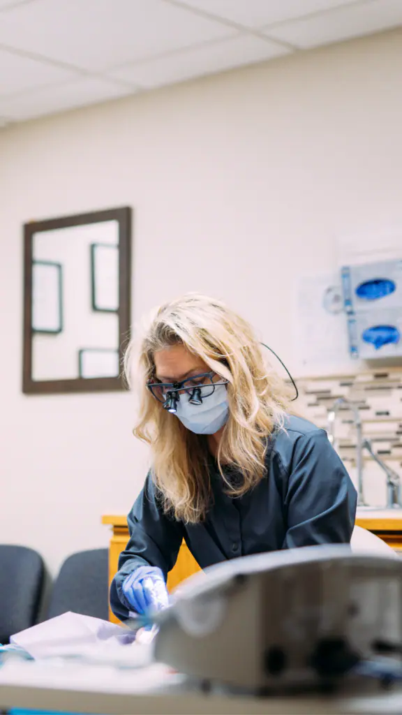 Female dental professional wearing a mask and magnifying loupes, focused on a patient during a procedure in a clinical setting.