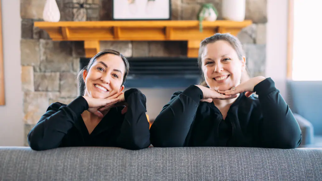 Two women lean on a sofa, resting their chins on clasped hands and smiling at the camera in a cozy living room with a stone fireplace behind them.