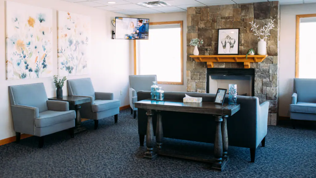 Cozy lounge with blue armchairs around a dark wooden table in a carpeted room with a stone fireplace and gallery art above it