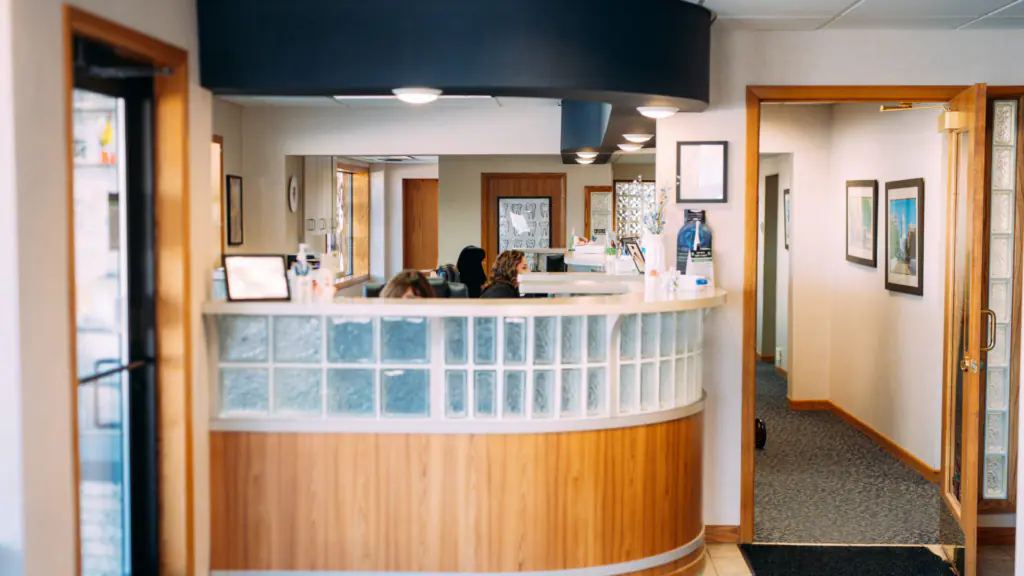 Dental clinic reception desk with curved wood counter and glass block facade, waiting area beyond, warm lighting and framed pictures on the wall